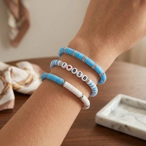 May include: A set of three stretch bracelets made of flat, circular clay beads. One bracelet is solid light blue with silver spacers, one features white beads with the text I heart BSB, and one is a mix of light blue and white beads with silver spacers.