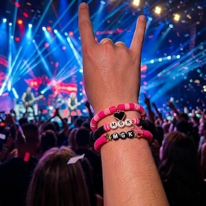 May include: A person's hand making a rock and roll hand gesture at a concert. The person is wearing two bracelets with the letters "MGK" on them. The bracelets are pink and black with star and heart charms. The background is a brightly lit stage.