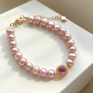 May include: A bracelet featuring a strand of round, light pink pearls with a gold-tone metal clasp and extender chain. A circular gold-tone charm set with a central round pink stone and a border of small clear crystals is attached to the strand.
