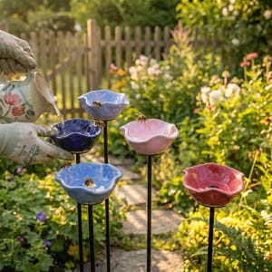 May include: A person pours water into a set of colorful ceramic bee water feeders in a garden. The feeders are in shades of blue, pink, and red, and are mounted on black metal stakes. The scene is set in a lush garden with flowers and greenery.