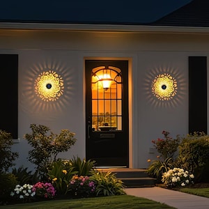 May include: Exterior view of a house at dusk, featuring two decorative solar lights with a sunburst design on the white wall. A black door with a glass panel is in the center, with a garden of flowers and bushes in front.