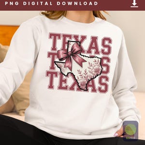 May include: White sweatshirt with the word "TEXAS" in maroon block letters. The state of Texas is depicted in the center, decorated with a pink bow and floral design. The sweatshirt is a casual style.