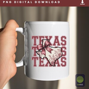 May include: White ceramic mug featuring the word "TEXAS" in maroon block letters. The state of Texas is illustrated in the center, adorned with floral designs and a maroon bow. The mug is held by a hand.