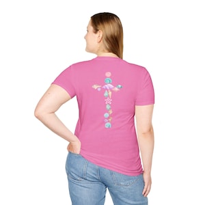 May include: A pink t-shirt featuring a cross design made of various seashells in pastel colors. The person is wearing blue jeans. The t-shirt is a crew neck style.