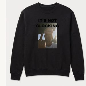 May include: Black sweatshirt featuring the text "IT'S NOT CLOCKING" above a photo of a person gesturing with their middle finger. The image is square and centrally placed on the garment.