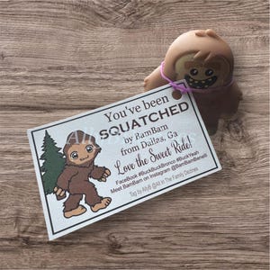 May include: A brown Bigfoot figurine with a tag that reads "You've been SQUATCHED by BamBam from Dallas, Ga." The tag also features a cartoon Bigfoot and a green tree. The figurine is attached to the tag with a purple string.