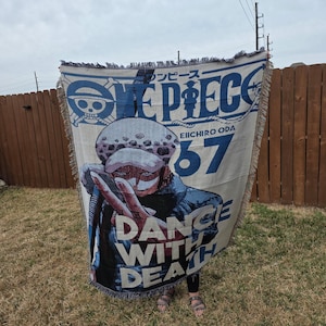 May include: A beige and blue woven throw blanket featuring the text "ONE PIECE" and an illustration of a character. The blanket also has the text "DANCE WITH DEATH" and the number "67". The blanket has a fringed edge.