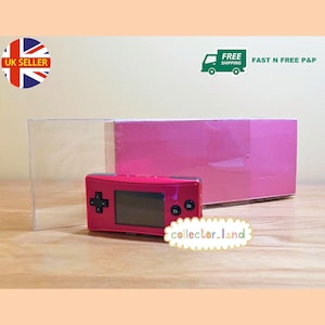 May include: A red handheld gaming console with a black screen and buttons, next to a pink box and a clear plastic case. The console has a directional pad and two buttons. The text "collector_land" is visible.