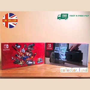 Puede incluir: Dos cajas de consola de juegos Nintendo Switch. Una caja es roja con un gráfico de Super Mario Odyssey. La otra muestra la consola y los mandos. Las cajas están sobre una superficie de madera.