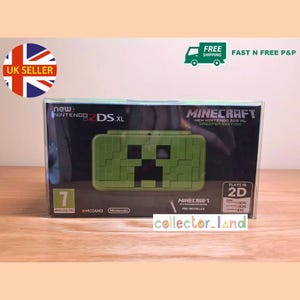 Peut inclure: Une nouvelle console de jeu Nintendo 2DS XL Creeper Edition. La console verte présente un design Creeper pixelisé de Minecraft. La boîte affiche le texte "Minecraft" et "Plays in 2D".