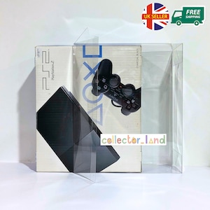 May include: A PlayStation 2 console in its original box, with a black controller visible. The box is encased in a clear plastic display case. The box has the PlayStation 2 logo and the words "CHARCOAL BLACK".