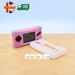 Könnte beinhalten: Eine rosa Handheld-Spielkonsole mit einem schwarzen Steuerkreuz und Tasten sowie einer separaten weißen Schutzhülle. Die Konsole hat einen Bildschirm und befindet sich auf einer Holzoberfläche. Der Text "collector_land" ist sichtbar.