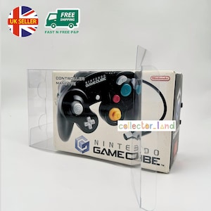 Puede incluir: Un mando negro de Nintendo GameCube en su embalaje original. El mando tiene botones coloridos y una cruceta. La caja presenta el logotipo de Nintendo GameCube y el texto "CONTROLLER MANETTE". El embalaje es de plástico transparente.