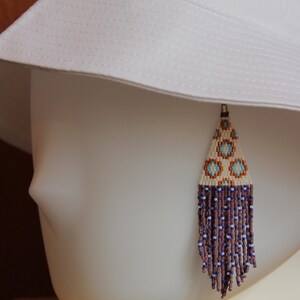 May include: A beaded earring featuring a triangular top section with a geometric pattern in cream, light blue, and copper, finished with a long fringe of dark purple and white beads, displayed on a white mannequin head wearing a white bucket hat.