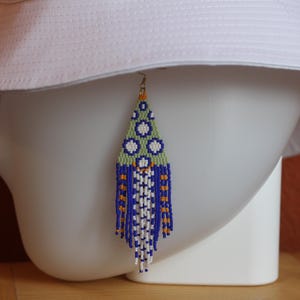 May include: A triangular beaded earring featuring a geometric pattern in shades of blue, light green, white, and orange. The top section displays a circular motif, while the lower portion consists of long, dangling fringe strands in matching colors.