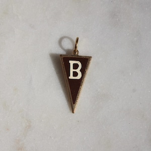 May include: A gold-coloured pendant in a pennant shape, featuring a dark brown background with a white letter "B". The pendant is suspended from a small loop, suggesting it's designed to be worn as jewellery.