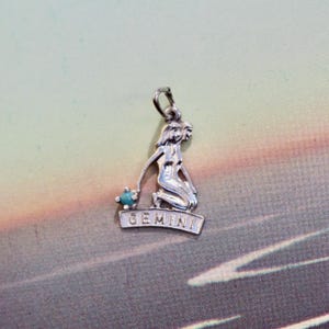 May include: Silver Gemini zodiac charm with a kneeling figure holding a turquoise bead. The word "GEMINI" is inscribed on a banner below the figure. The charm has a loop at the top for attaching to a necklace or bracelet.