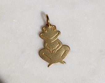 Può includere: Un ciondolo a forma di rana color oro con un anello nella parte superiore per attaccarlo a una collana o un braccialetto. La rana è in posizione seduta con le braccia incrociate. Il ciondolo ha una finitura liscia e lucida.