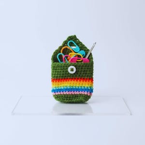 May include: A small, green crocheted pouch with a rainbow stripe design, holding colourful stitch markers and a crochet hook. The pouch is secured with a white button. A craft accessory for crocheters.