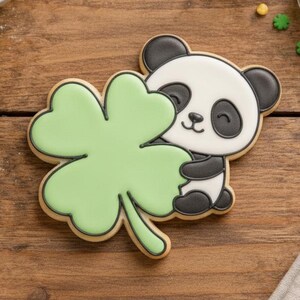 May include: A decorated sugar cookie in the shape of a panda hugging a green shamrock. The panda is black and white with a smiling face. The shamrock is light green with a darker green outline. The cookie is on a wooden surface with other baking supplies.