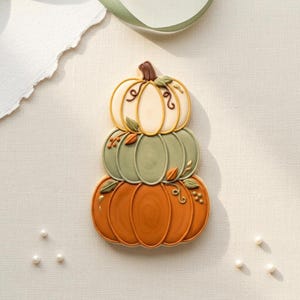 Peut inclure: Une pile de trois biscuits décorés en forme de citrouilles. La citrouille du haut est crème, celle du milieu est vert sauge et celle du bas est orange. Chaque citrouille présente des détails en glaçage, dont des feuilles, des vrilles et des baies marron.