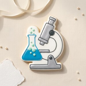 May include: A sugar cookie decorated with white icing, featuring a microscope and a flask with blue liquid and green bubbles. The cookie has gray and blue accents and is on a light beige surface.