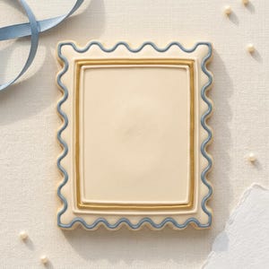 May include: A rectangular biscuit with a cream-coloured centre and a gold and blue border. The biscuit has a scalloped edge and is decorated with a blue ribbon and pearls.
