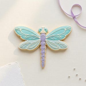 May include: A dragonfly-shaped cookie with light blue and white icing details. The wings are a gradient of light blue, with white decorative lines. The body is purple, and the cookie is on a white surface.