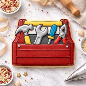 May include: A sugar biscuit shaped like a red toolbox with a yellow handle. The biscuit features icing details of a silver hammer with a yellow handle, a silver wrench, a blue and yellow screwdriver, and silver pliers with red handles.