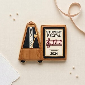 May include: A cookie shaped like a metronome and a rectangular sign. The metronome is brown with a black face and a pendulum. The sign has a cream background with black text reading STUDENT RECITAL, a red musical staff, and the year 2024 in black.