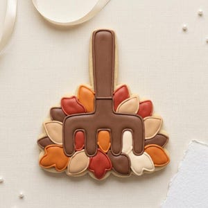 May include: A sugar biscuit shaped like a garden rake resting on a pile of autumn leaves. The rake is dark brown, while the leaves are decorated in shades of orange, red, cream, and brown icing. The design features a smooth, matte finish.