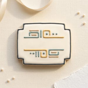 Peut inclure: Un biscuit rectangulaire avec une base blanche et une bordure noire. Le biscuit présente des motifs abstraits en glaçage jaune, bleu et marron. Le biscuit est décoré de points et de lignes, et est présenté sur une surface crème.