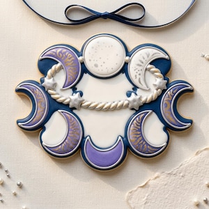 Peut inclure: Un biscuit décoratif en forme d'arrangement céleste comprenant une pleine lune centrale, plusieurs croissants de lune dans des tons de violet et de blanc, et une bordure argentée en forme de corde avec des étoiles, sur un fond crème.