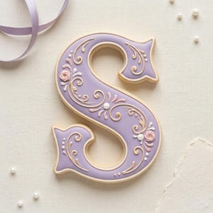 May include: A letter S shaped biscuit decorated with smooth lavender icing, featuring intricate cream-coloured scrollwork, small white piped dots, and two delicate pink icing roses. The biscuit has a clean, defined edge and a flat surface.