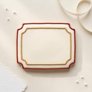 May include: A rectangular, off-white biscuit with a decorative red and gold border. The biscuit has a scalloped edge and is surrounded by a cream-coloured background with a white ribbon and small pearls.