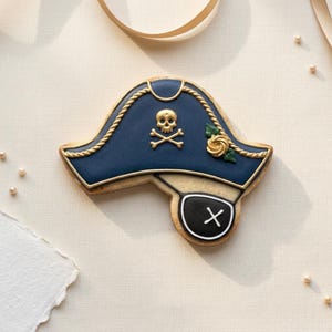 May include: A decorated sugar cookie shaped like a navy blue pirate hat with gold rope trim, a gold skull and crossbones emblem, and a small gold rose. Attached to the hat is a black eye patch cookie featuring a white X symbol in the center.