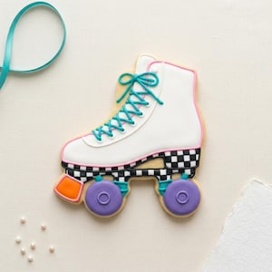 May include: A roller skate cookie with a white boot, pink and black checkered trim, and purple wheels. The laces are teal, and the cookie has an orange accent. The background is a light beige.