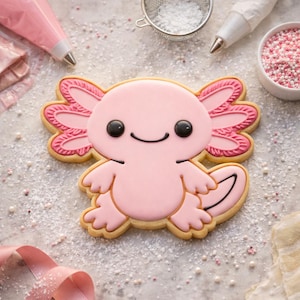 May include: A decorated sugar cookie in the shape of a pink axolotl with black eyes and a smiling mouth. The cookie is surrounded by baking supplies, including piping bags, sprinkles, and a sieve.