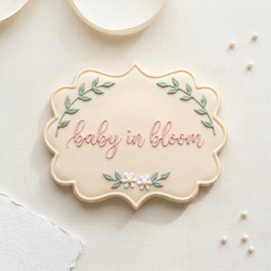 May include: A light beige cookie with a scalloped edge, decorated with the phrase "baby in bloom" in pink script. The cookie is adorned with green leaf sprigs and small white and yellow flowers, suggesting a baby shower theme.