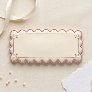 May include: A rectangular biscuit with a scalloped edge, decorated with a light pink border and gold accents. Small floral designs are placed at each corner. A white ribbon and pearls are in the background.