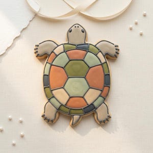 May include: A turtle-shaped biscuit with a colourful, geometric shell design. The shell features orange, green, and beige segments outlined in dark grey. The biscuit has a light beige body and is set against a neutral background.