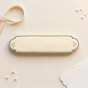 Peut inclure: Un biscuit rectangulaire blanc cassé avec une bordure bleu clair et des accents dorés. Le biscuit a des extrémités arrondies et trois points dorés à chaque extrémité. Un ruban crème et des perles éparses sont en arrière-plan.