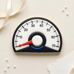 May include: A cookie shaped like a speedometer, with a white face, black border, and a red needle. The speedometer reads 0 to 70, with numbers and hash marks. The cookie has a black center and a blue accent at the bottom.
