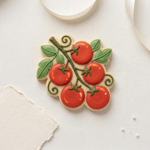 May include: A decorated sugar biscuit shaped like a cluster of red tomatoes on a green vine with green leaves. The biscuit has a light tan base and is decorated with icing.