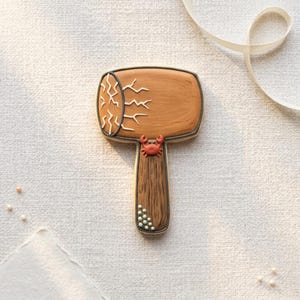 May include: A decorative cookie shaped like a wooden mallet, featuring a brown head with white crack detailing and a brown handle. A small red crab is placed on the handle. The cookie is outlined in a darker brown.