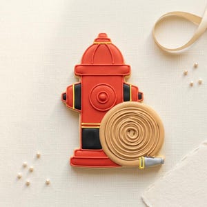 May include: A decorated cookie in the shape of a red fire hydrant with black and yellow accents. A tan-colored rolled-up hose is attached to the hydrant. The cookie is on a white surface with a ribbon and small pearls.