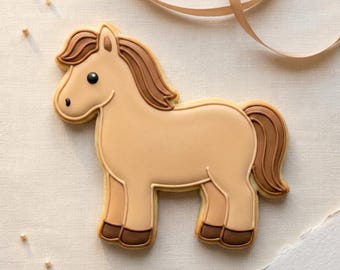 May include: A horse-shaped cookie with tan icing and brown details for the mane, tail, and hooves. The cookie has a smiling face and is decorated with a light brown outline. A beige ribbon and small white sprinkles are in the background.