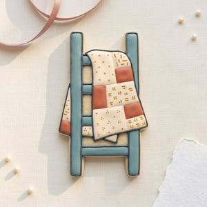 May include: A decorated sugar cookie in the shape of a bed with a quilt. The bed frame is light blue, and the quilt is cream, red, and gold. The quilt has a patchwork design with dots and dashes.