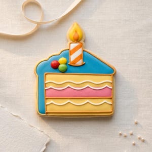 May include: A birthday cake-shaped biscuit with blue icing, yellow and pink layers, and a lit candle. The biscuit is decorated with red, yellow, and green sweets. A white ribbon and pearls are in the background.