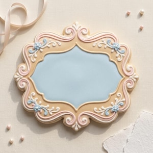 May include: A decorative cookie with a light blue centre and a beige frame. The frame is adorned with pink, white, and light blue floral and scroll designs. The cookie is on a cream-coloured surface.